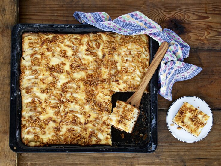 Apfelkuchen vom Blech mit Krokant, angeschnitten auf dem schwarzen Ofenblech serviert. 