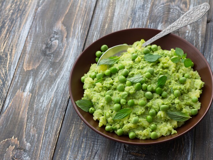 Draufsicht: Ein Teller mit Mushy Peas, also pürierten Erbsen, steht auf einen Holztisch. In dem Püree ist ein Löffel aus Metall.