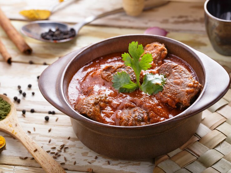 Nur ein Topf, aber viel Geschmack und raffinierte Aromen: Das asiatische Lammcurry Rogan Josh! Draufsicht, schräg: Ein Tontopf mit dem asiatischen Currygericht Rogan Josh steht auf einer Bastmatte, die auf einem Holztisch liegt. Drumherum liegen Holzlöffel mit Gewürzen.
