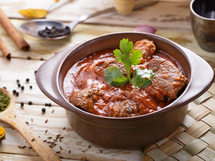 Draufsicht, schräg: Ein Tontopf mit dem asiatischen Currygericht Rogan Josh steht auf einer Bastmatte, die auf einem Holztisch liegt. Drumherum liegen Holzlöffel mit Gewürzen.