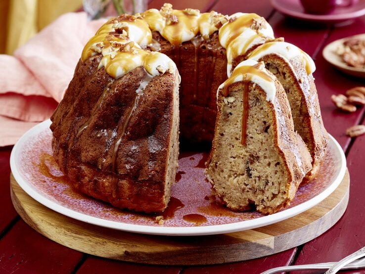Eine Kuchenplatte mit dem angeschnittenen Apfel-Bananen-Gugelhupf mit Karamell-Soße übergossen und Pekan-Nüssen überstreut vor einem dunkel-roten Hintergrund.