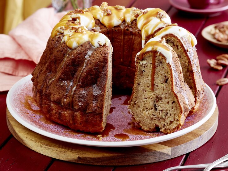 Eine Kuchenplatte mit dem angeschnittenen Apfel-Bananen-Gugelhupf mit Karamell-Soße übergossen und Pekan-Nüssen überstreut vor einem dunkel-roten Hintergrund.