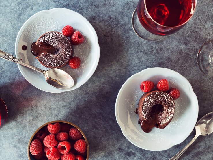 Schoko-Lavakuchen von oben auf weißen Tellern mit Himbeeren und Puderzucker auf einem grauen Tisch neben frischen Himbeeren und Gläsern mit Rotwein. 