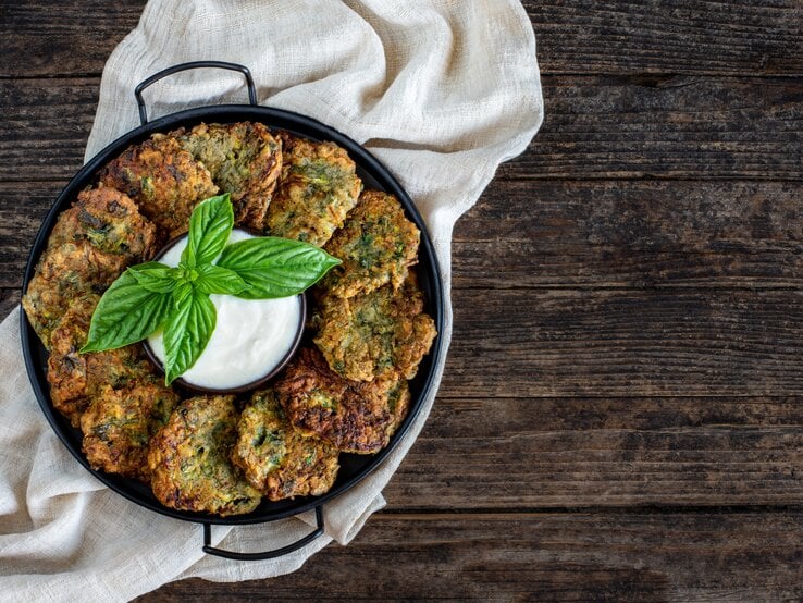 Reiche die Brokkoli-Kartoffelpuffer mit einem schnell angerührten Dip. Eine Pfanne mit Brokkoli-Kartoffelpuffern, die im Kreis angeordnet sind, in der Mitte eine Schale mit Dip. Draufsicht.