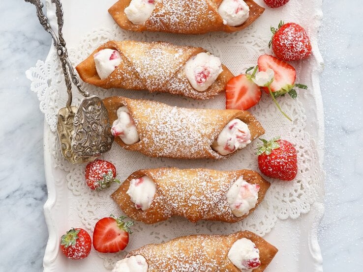 Cannoli mit Ricotta-Erdbeer-Füllung Italienische Cannoli mit Ricota-Erdbeer-Füllung, bestäubt mit Puderzucker und serviert auf einem weißen, verzierten Kuchenteller, angereicht mit frischen Erdbeeren.