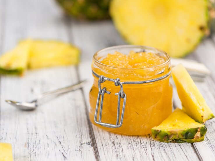 Ein Glas Ananas-Marmelade steht auf einem hellen Dielenboden. Drum herum liegen dreieickige Ananasstücke und ein Löffel. Im Hintergrund eine angeschnittene Ananas.