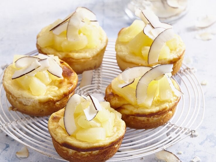 Auf einem Kuchengitter stehen mehrere Pina-Colada-Mini-Käsekuchen. Sie sind mit Kokosraspeln verfeinert. 