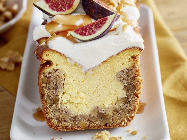 Angeschnittener Feigen-Walnuss-Kuchen auf weißem Teller, im unscharfen Hintergrund Walnüsse und ein gelbes Küchentuch. 