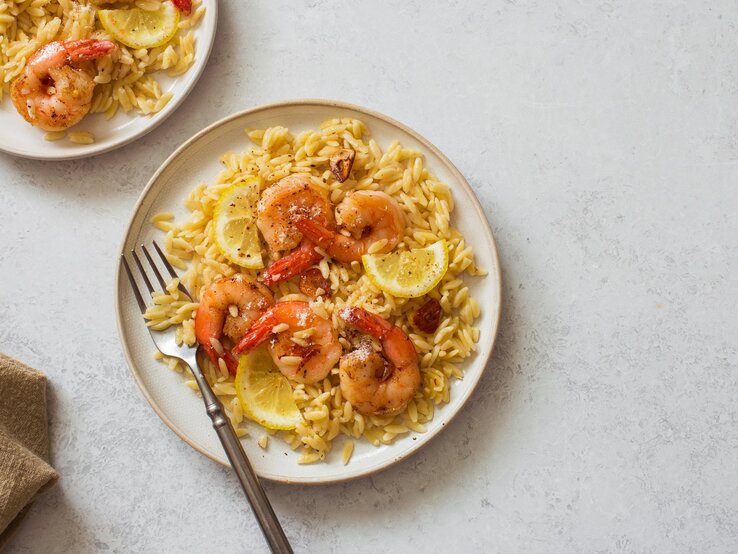 Draufsicht: Zwei Teller mit Orzo-Pasta, Zitronenscheiben und Garnelen. Auf einem Teller liegt eine Gabel.