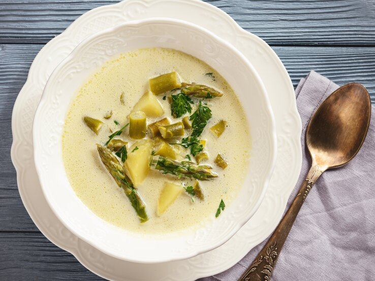 Spargel-Käse-Süppchen in verspielter Schüssel auf blauem Holz. 
