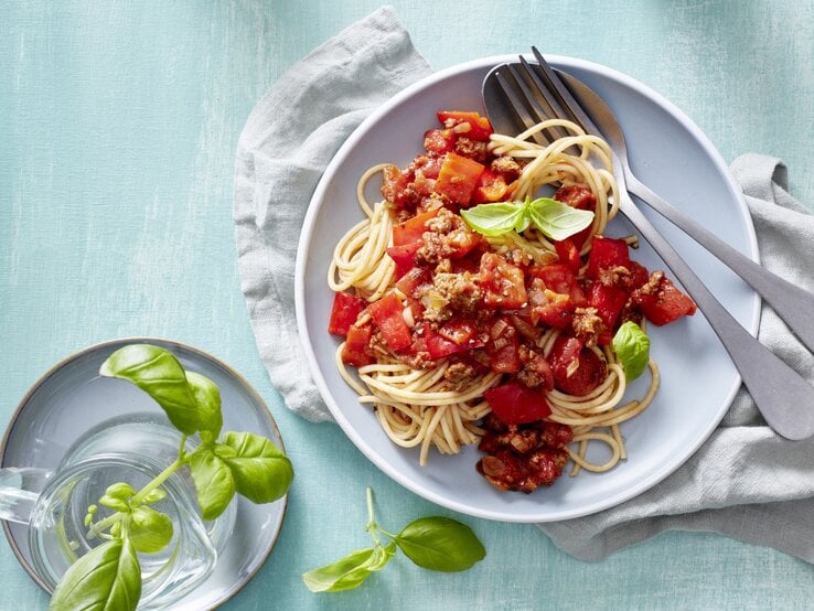 Pasta mit Hack und Paprika, garniert mit frischem Basilikum und serviert auf einem Teller auf hellblauem Tisch. 