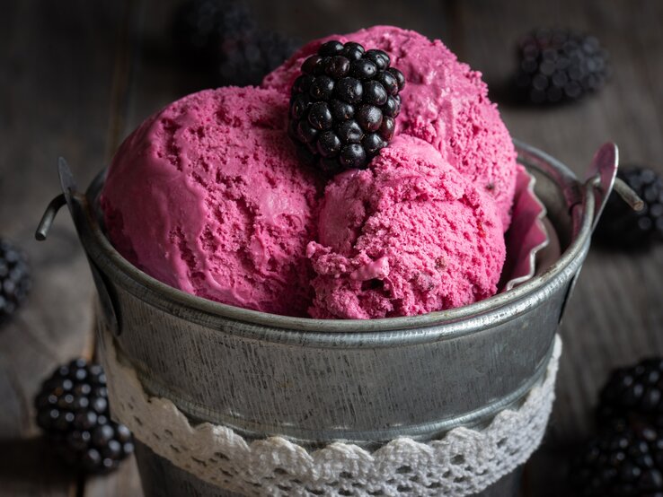 Zum Garnieren vom Brombeer-Bananen-Eis eignen sich ein paar frische Beeren. Ein kleiner Metalleimer mit 3 Kugeln Brombeer-Bananen-Eis, garniert mit einer Brombeere, dunkle Umgebung.