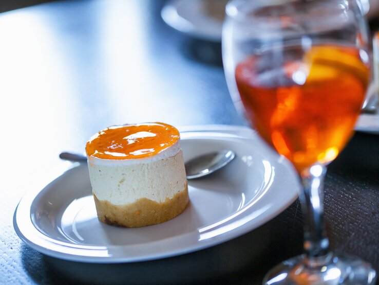 Ein kleiner Aperol-Käsekuchen mit einem orangenen Guss aus Orangensaft, Prosecco und Aperol neben einem Glas mit Aperol Spitz | © Shutterstock/ NatashaPhoto