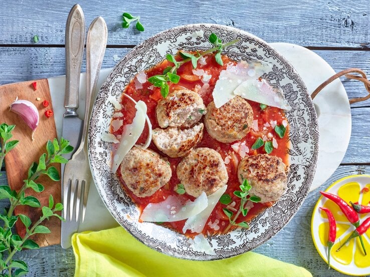 Hackbällchen mit Ricotta und Oregano, serviert auf einem Sud aus Tomaten, garniert mit frischen Kräutern und groben Parmesanspänen auf einem traditionell italienisch dekorierten Teller, platziert auf einem hellen Holztisch. 