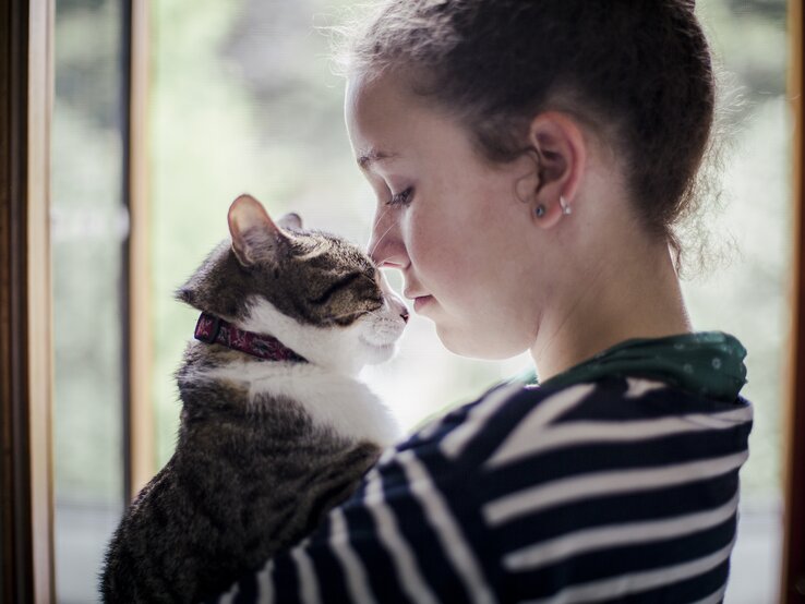 Eine junge Frau steht am Fenster und hält ihre Katze im Arm. Sie schauen sich an. 