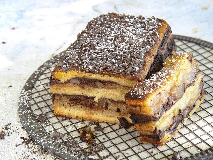 Croque Cake mit Apfel-Birnen-Schokolade, bestehend aus mehreren Schichten Kastenkuchen, getoppt mit Schichten aus Birne und Schokoladencreme und bestreut mit Puderzucker und Schokostreuseln. 