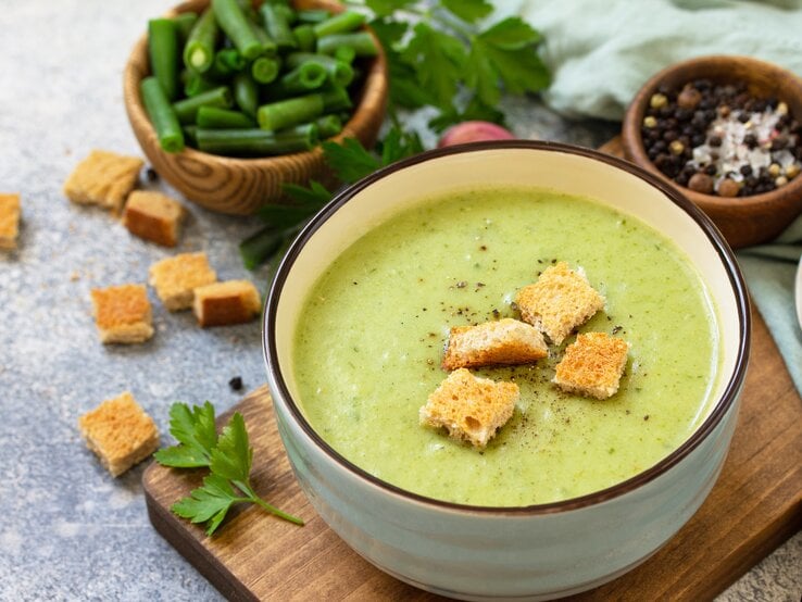 Die mögen alle: Eine gute Bohnensuppe mit Schmand! Draufsicht: Eine Schüssel mit einer grünen cremigen Flüssigkeit steht auf einem Holzbrettchen. Auf den Flüssigkeit liegen brotstücke. Neben der Schüssel liegen Petersilie sowie einige Gewürze.