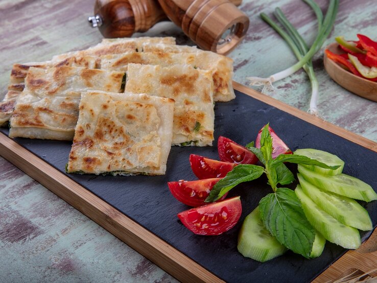 Gözleme mit Spinat-Schafskäsefüllung auf Brett mit Salat. Daneben Lauch und Gewürzmühlen aus Holz. In eienr Schale Paprikastreifen. Draufsicht.