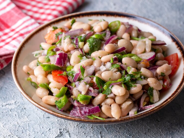 Piyaz auf einem Teller mit Bohnen und Gemüse auf einem rauen Untergrund. Neben dem Teller liegt ein Tuch.