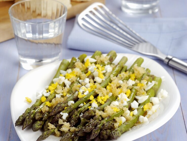 Grüner Spargel auf polnische Art, serviert mit weich gekochtem Ei und einer feinen Estragon-Vinaigrette. Alles auf weißem Teller und violettem Holztisch.