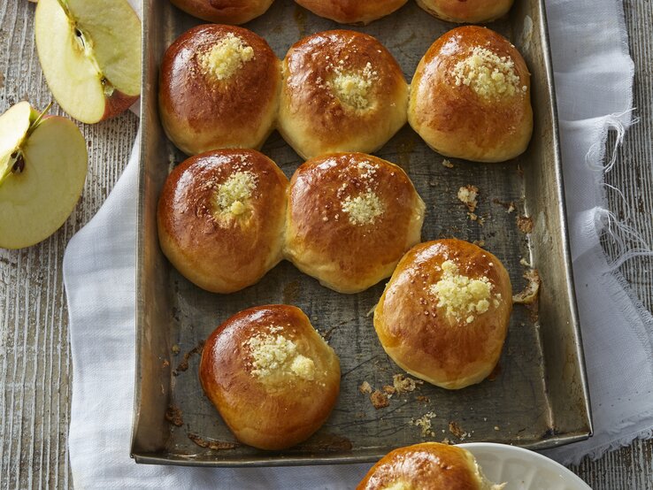 Auf einem Backblech liegen einige Hefebrötchen mit Apfel. Sie sehen fluffig und weich aus. Daneben sind aufgeschnittene Äpfel zu sehen. 
