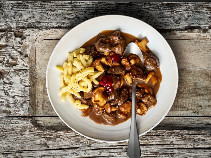 Ein weißer Teller mit einer Portion des Rehgulaschs mit Spätzle und Preiselbeeren, verfeinert mit Pfifferlingen. Ein Löffel ist auf dem Teller platziert, der vor einem Holzhintergrund von oben dargestellt ist.  