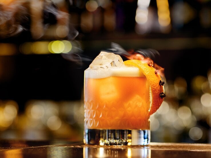 Ein Glas Whisky Sour steht auf einem Bartresen. Im Hintergrund verschwimmen einige Lichter der Bar. Das Glas ist angerichtet mit einer Orangenscheibe. Im Cocktail schwimmt ein Eiswürfel.