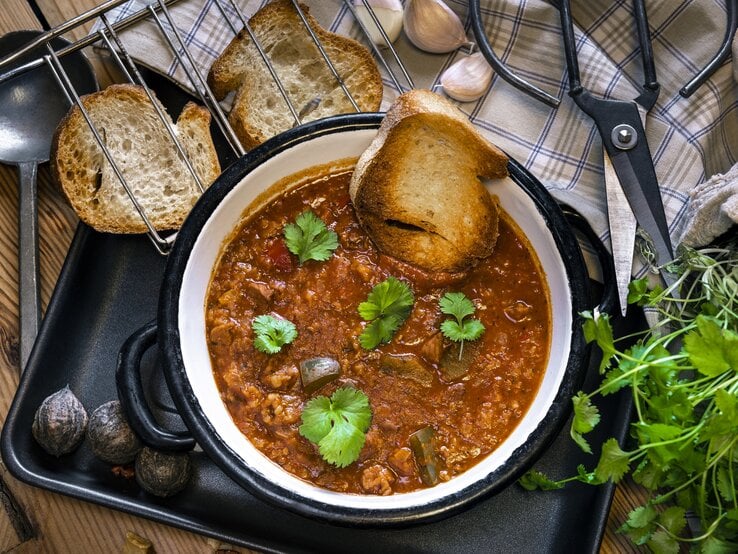 Auf einem Holztisch liegen Brotschreiben, eine Schere, frische Kräuter und ein Suppenlöffel. Darüber hinaus steht dort ein Topf Lammsuppe aus Georgien. Das Gericht nennt man Kharcho.