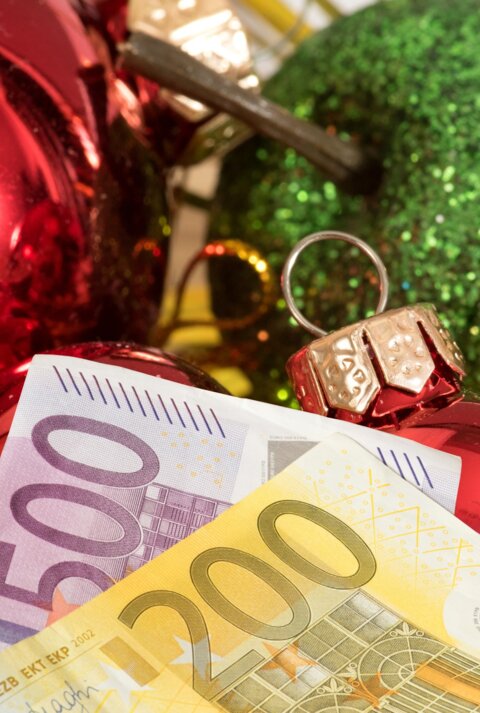 Ein 200-Euro- und ein 500-Euro-Schein liegen vor Weihnachtsdeko. Im Hintergrund sind ein grüner Deko-Apfel und Christbaumkugeln zu sehen.