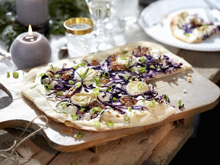 Ein Rotkohl-Flammkuchen mit Ziegenkäse liegt auf einem Holzbrett auf einem Holztisch. Im Hintergrund stehen Gläser und mehrere Kerzen. Der Flammkuchen ist angerichtet mit Walnüssen und Frühlingszwiebelringen.