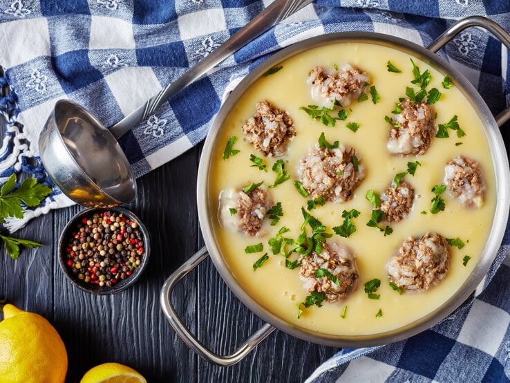 Griechische Hackbällchensuppe mit Zitrone in einem Topg aus Edelstahl neben einer Suppenkelle, Pfefferkörnern und frischer Zitrone auf einem blauen Holztisch mit blau karierter Tischdecke.
