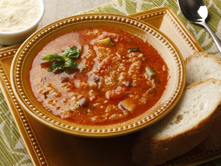 Ein Teller Couscous-Suppe mit Zucchini und Tomate steht auf einem Tablett. Daneben lieben zwei Scheiben Brot.