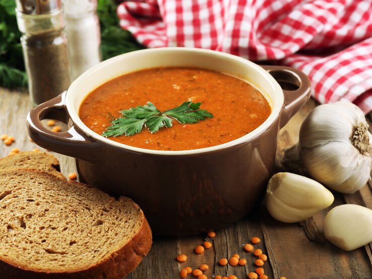 Frontalansicht: Auf einem Tisch steht eine Schale mit einer roten Suppe, die mit einem Blatt Petersilie garniert ist. Drumherum liegen Knoblauchzehen, eine Scheibe Brot und trockene rote Linsen.