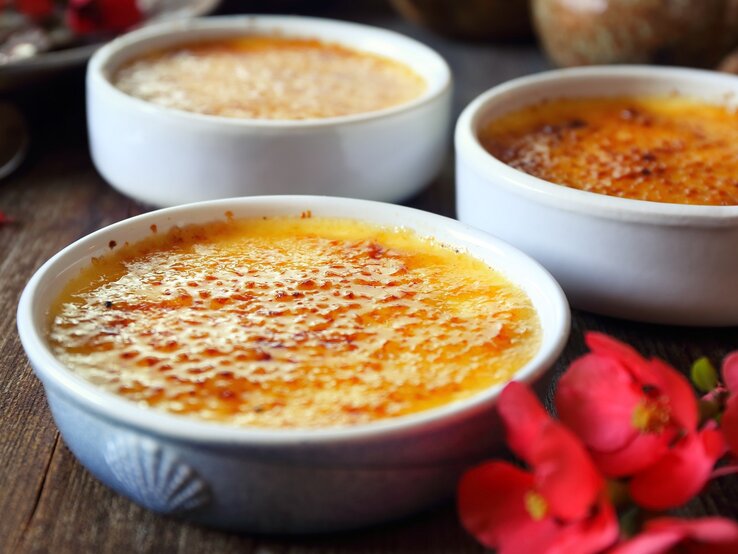 Drei Eierlikör-Crème brûlée Schalen auf einem dunklen holztisch mit roten Blüten, seitlich fotografiert.