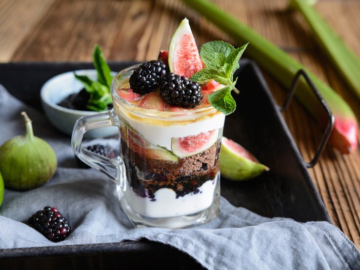 Schichtdessert mit Rhabarber, Feigen, Ricottacreme, Kaffeelikörkeksen Brombeeren und Minzblatt auf schwarzem Tablette mit Beeren und Feigen. Daneben auf Holztische Rhabarber. Frontalansicht.