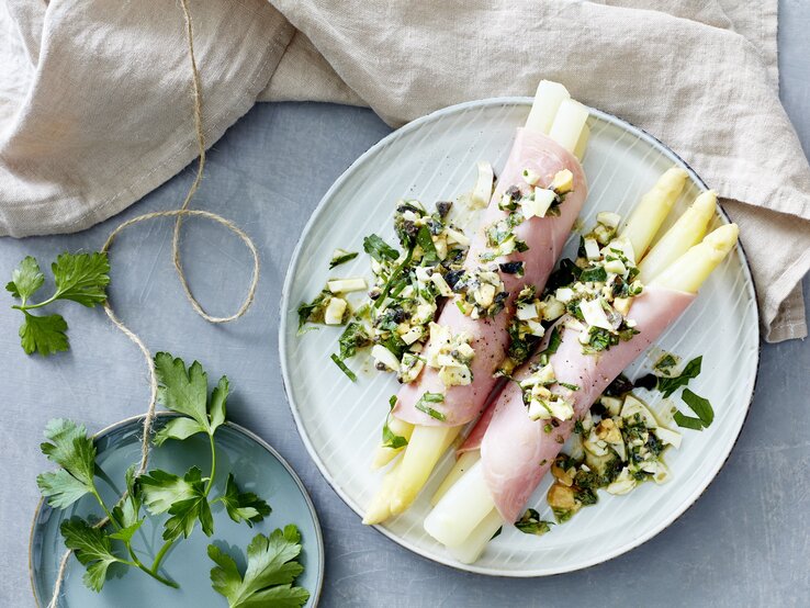 Mediterraner Spargel eingewickelt in Schinken auf einem hellblauen Teller neben Petersilie auf einem gedeckten hellblauen Tisch