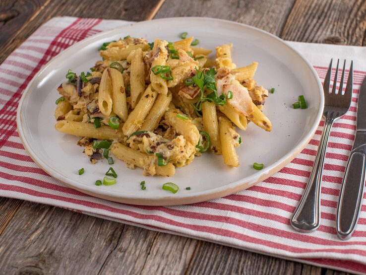 Ein weißer Teller mit den Eier-Schinkennudeln auf einem rustikalen Holztisch mit rot-weiß-karierter Tischdecke.