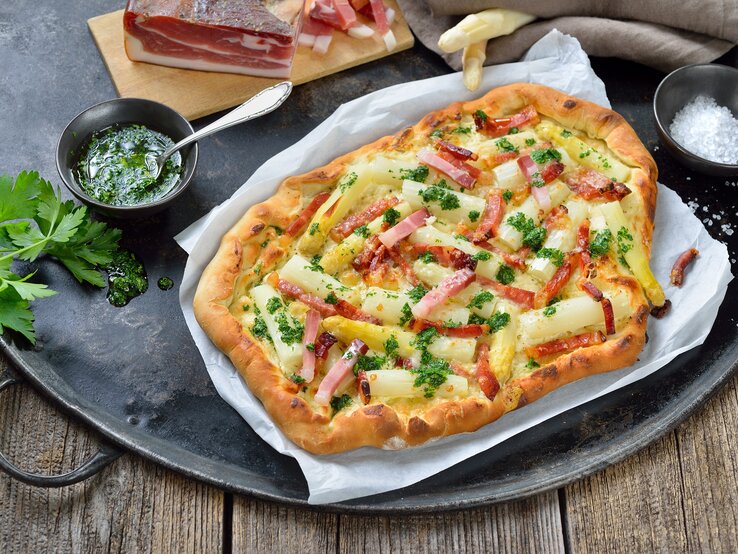 Spargel-Flammkuchen auf einem Stück Backpapier. Daneben verschiedene Zutaten.