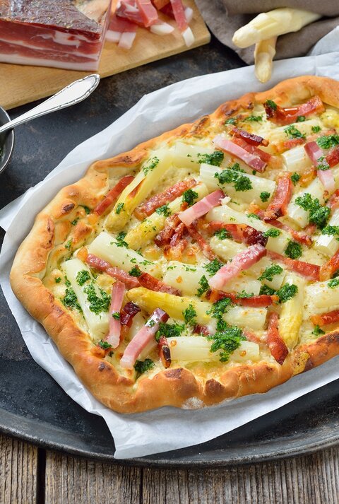 Spargel-Flammkuchen auf einem Stück Backpapier. Daneben verschiedene Zutaten.