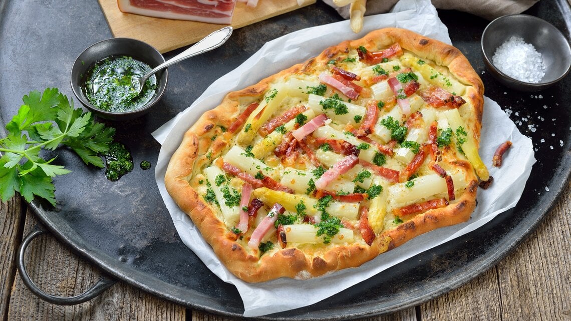 Spargel-Flammkuchen auf einem Stück Backpapier. Daneben verschiedene Zutaten.