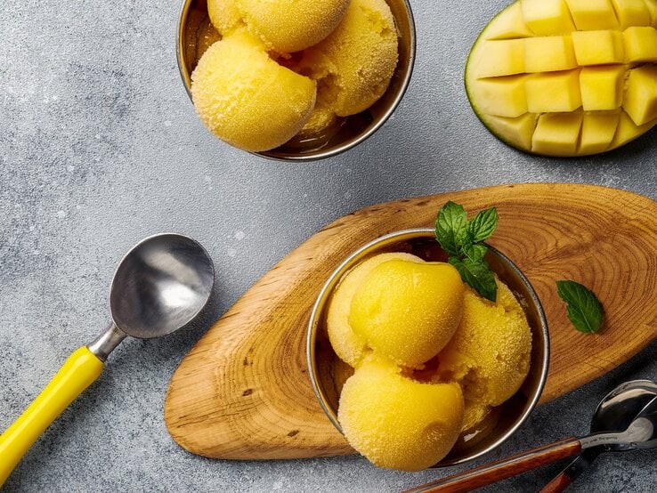 Ein paar Kugeln Mango-Joghurt-Eis in zwei Schälchen auf einem grauen Tisch. Daneben eine eingeritzte Mangohälfte und ein paar Löffel. Alles direkt von oben fotografiert.