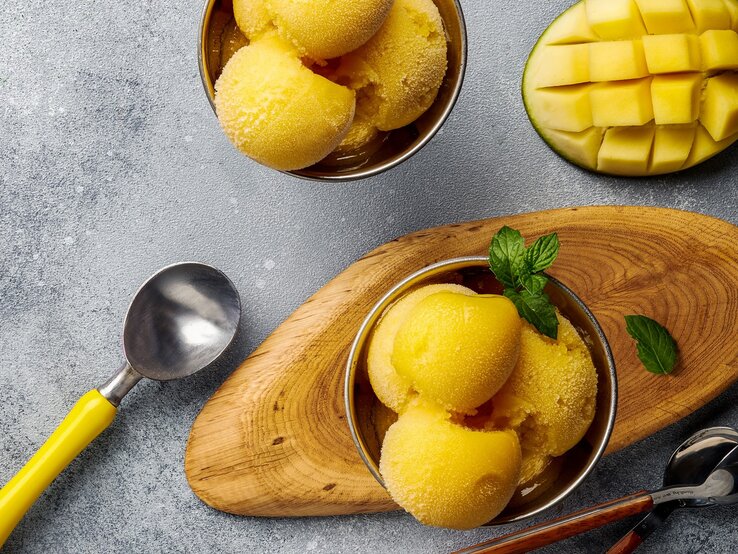 Mango-Joghurt-Eis Ein paar Kugeln Mango-Joghurt-Eis in zwei Schälchen auf einem grauen Tisch. Daneben eine eingeritzte Mangohälfte und ein paar Löffel. Alles direkt von oben fotografiert.