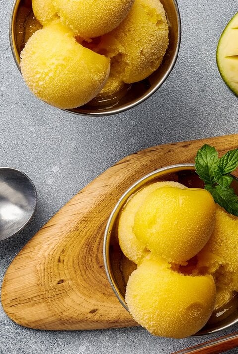 Ein paar Kugeln Mango-Joghurt-Eis in zwei Schälchen auf einem grauen Tisch. Daneben eine eingeritzte Mangohälfte und ein paar Löffel. Alles direkt von oben fotografiert.