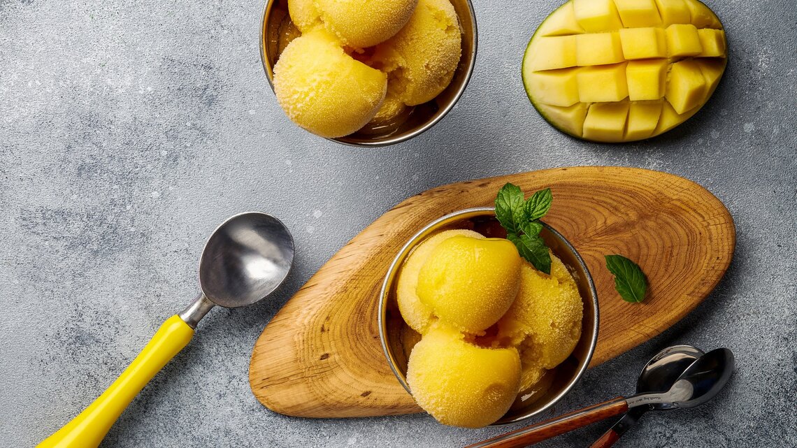 Ein paar Kugeln Mango-Joghurt-Eis in zwei Schälchen auf einem grauen Tisch. Daneben eine eingeritzte Mangohälfte und ein paar Löffel. Alles direkt von oben fotografiert.