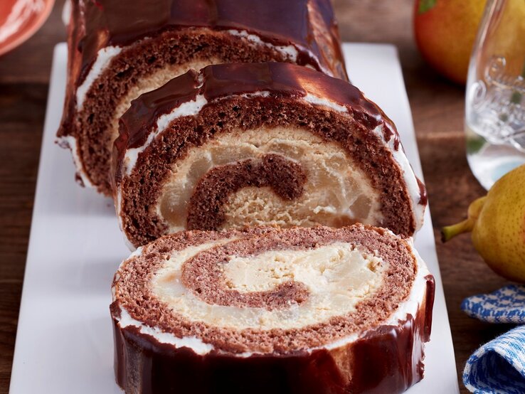 Eine lange weiße Kuchenblatte auf der die angeschnittene Schokorolle mit Mascarpone-Creme und Birnen zu sehen ist, mit einem dunklen Schokoladen-Guss bedeckt,neben Birnen, Blumen und einem Teller mit Gabeln vor einem braun-blauen Hintergrund.