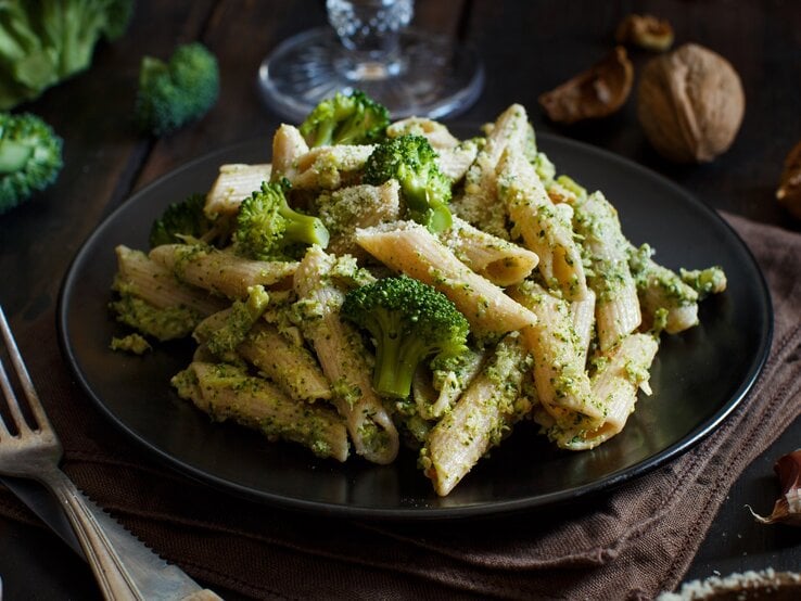 Ein Teller Brokkoli-Grünkohl-Pasta, daneben eine Gabel, im Hintergrund Walnüsse und Brokkoli.
