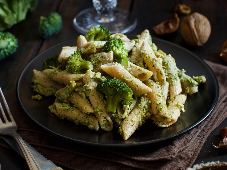 Ein Teller Brokkoli-Grünkohl-Pasta, daneben eine Gabel, im Hintergrund Walnüsse und Brokkoli.