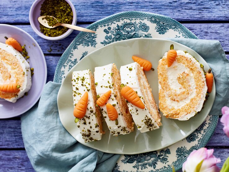 Mehrere Stücke Rübli-Zitronenbiskuitrolle mit süßer Mini-Karotten-Deko auf einem Teller vor lila Hintergrund. 