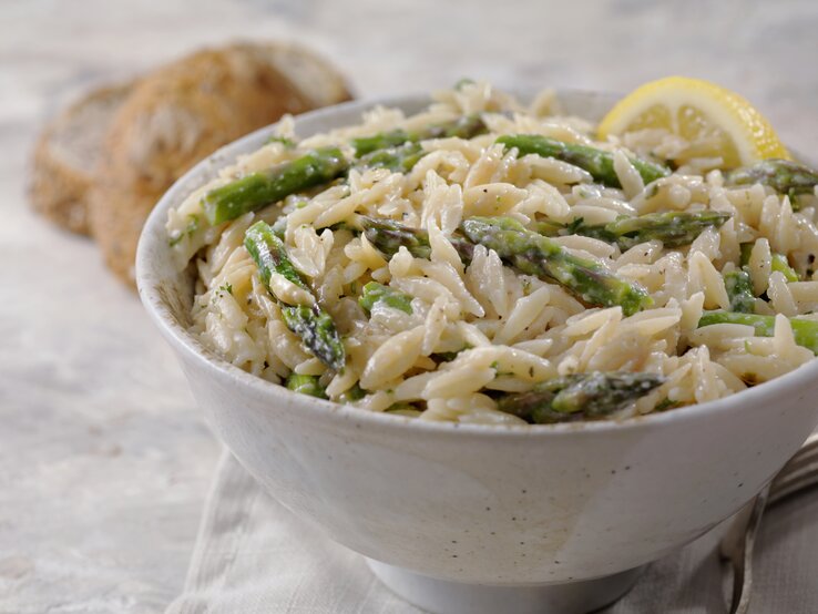 Im Vordergrund steht eine weiße Schüssel mit einem cremigen Spargel-Orzo-Topf mit Parmesan. Daneben liegen zwei Brötchen.