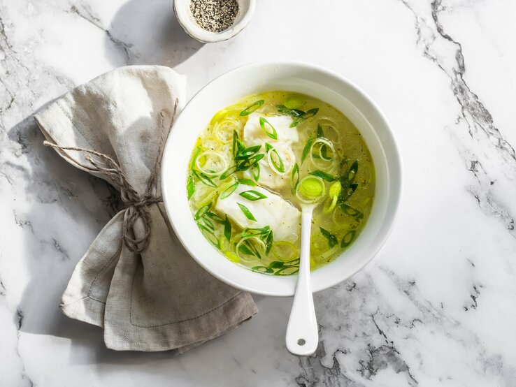 Draufsicht: eine helle Schale mit einer Leichte Lauch-Kabeljau-Suppe steht auf einen Untergrund aus hellem Marmor. In der Suppe ist ein Suppenlöffel.